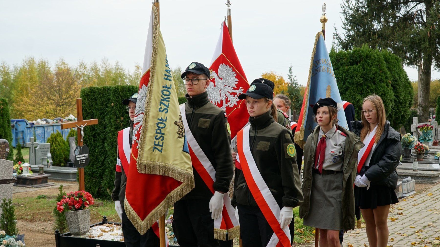 Poczet sztandardowy uczniów ubrany w odświętne mundury, mundury harcerskie i ubrania galowe stoi przy grobie.