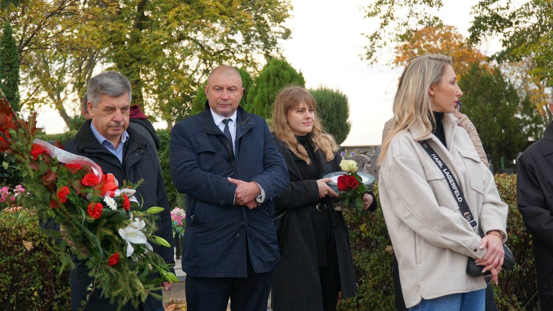 Gruba osób, która zebrała się wspólnie nad grobem. Mężczyzna po lewej trzyma w rękach wieniec z czerwonymi różami i wstęgą. Kobieta po prawej trzyma w ręku trzy róże i znicz. Mężczyzna w środku stoi pomiędzy nimi i przygląda się nagrobkowi.