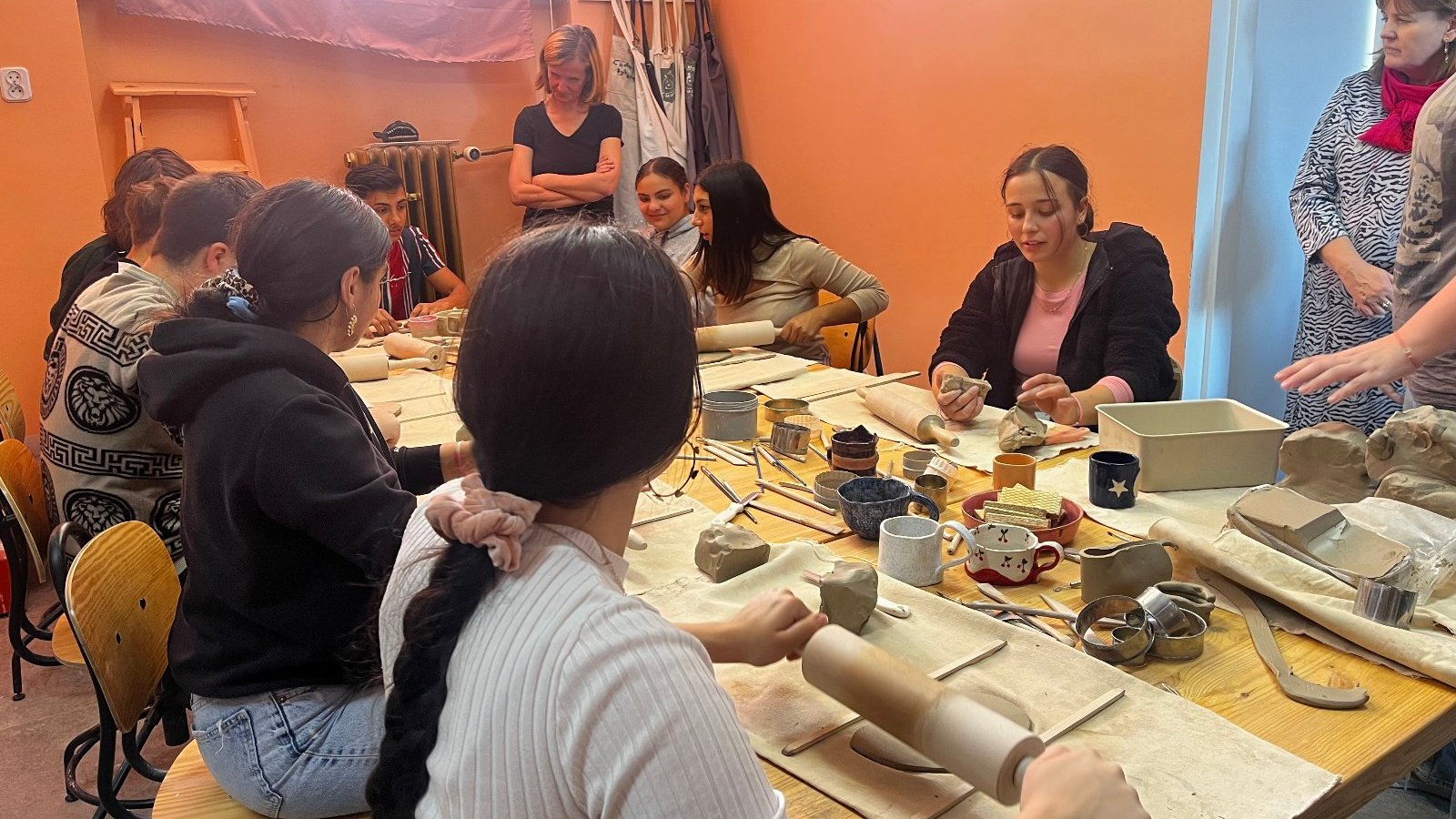 Na zdjęciu widoczni są uczniowie, którzy wytwarzają wyroby ceramiczne z gliny, widoczne są również nauczycielki, prowadzące zajęcia, a na stole materiały potrzebne do pracy na przykład glina, woda