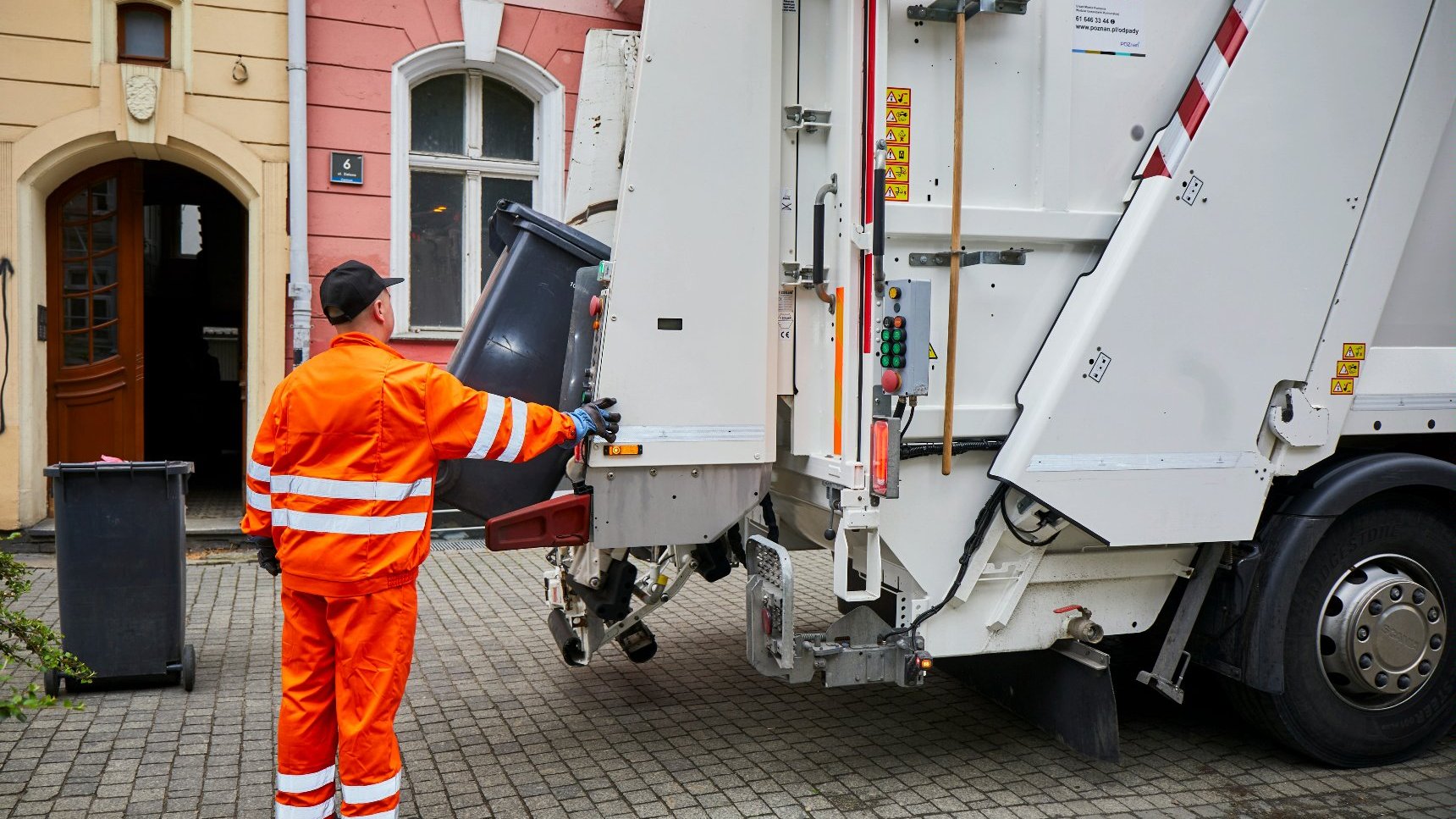 Moment odbioru odpadów komunalnych: pracownika obsługującego śmieciarkę i moment opróżniania czarnego pojemnika na odpady zmieszane.