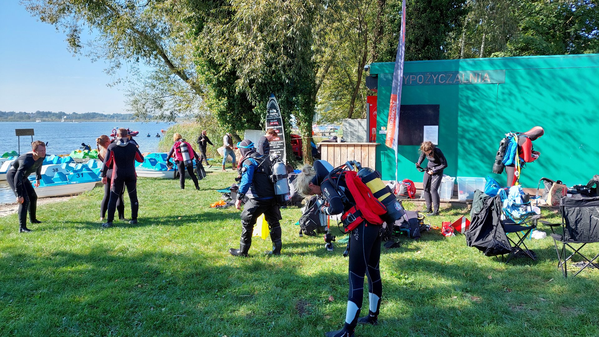 Na zdjęciu widać kilkanaście osób zakładających kombinezony rnurkowe. W tle wysokie drzewa. Po lewej stronie widoczny fragment jeziora z plażą. Przy brzegu kilka łódek typu opimist - grafika artykułu