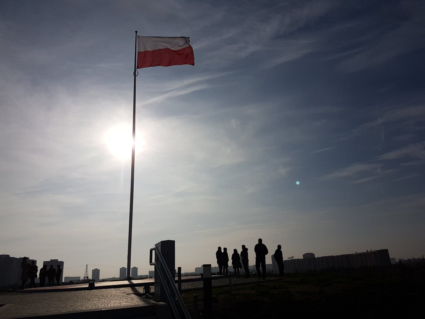 Popołudnie. Na szczycie Kopca stoi wysoki maszt z flagą Polski. Pod masztem stoją ludzie i oglądają widoki. W tle panorama Poznania. - grafika artykułu