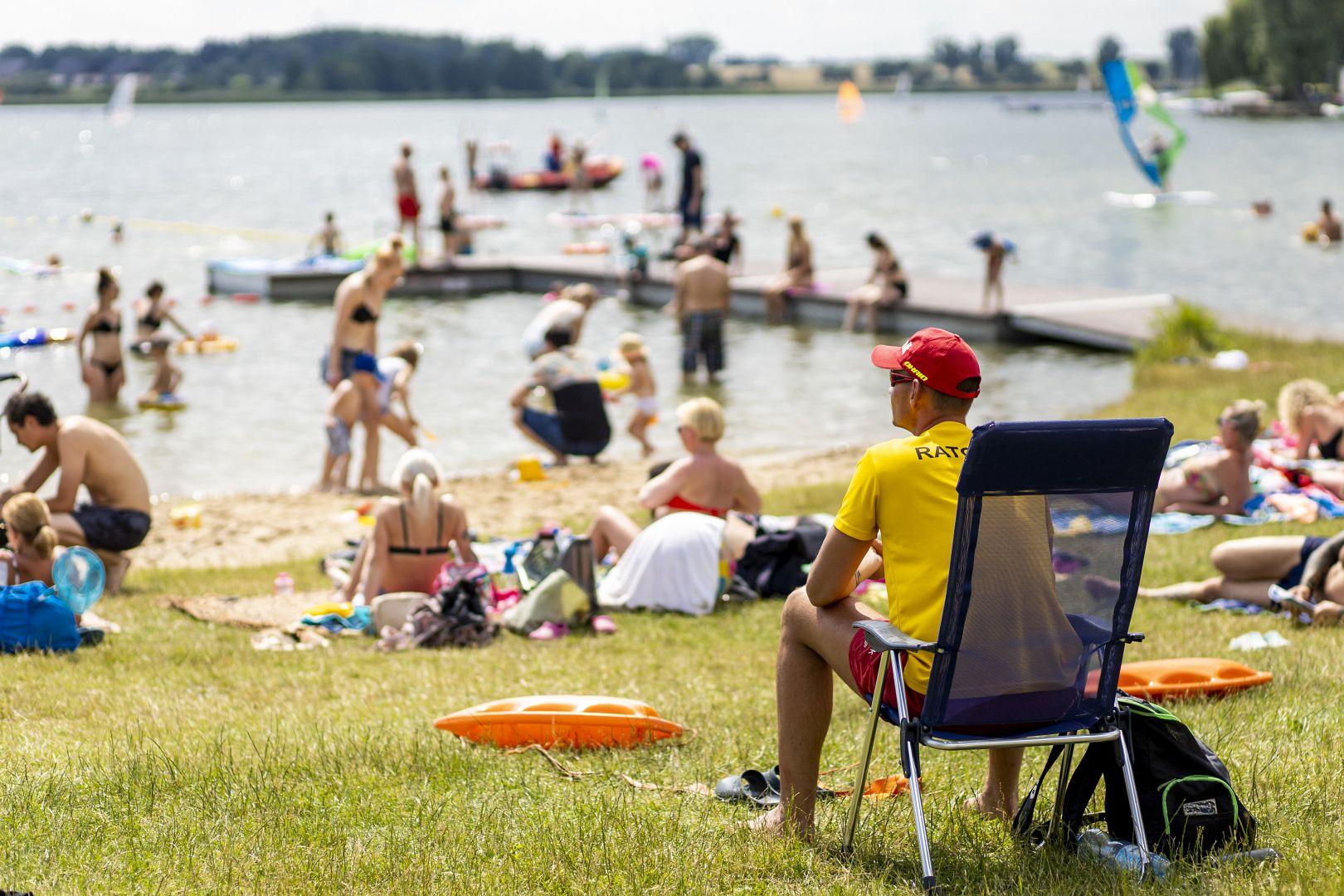 Na pierwszym planie mężczyzna siedzi na krześle letniskowym. Jest ubrany w żółtą koszulkę z napisem "Ratownik", czerwoną czapkę i kąpielówki. Obserwuje, co dzieje się w jeziorze. Wokół niego ludzie leżą na plaży lub kąpią się w jeziorze. - grafika artykułu