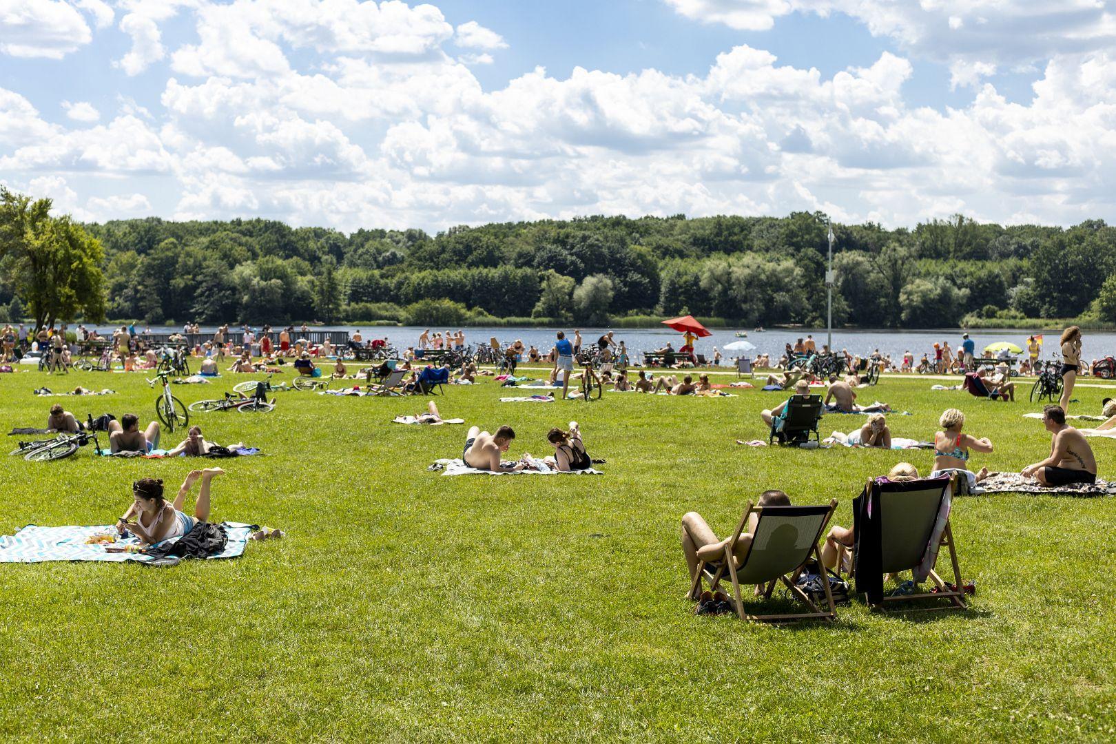 Na zielonej trawie różni ludzie leżą na kocach lub siedzą na leżakach. W oddali jezioro, w którym pływają inne osoby. W tle las i niebieskie niebo pokryte chmurami. - grafika artykułu