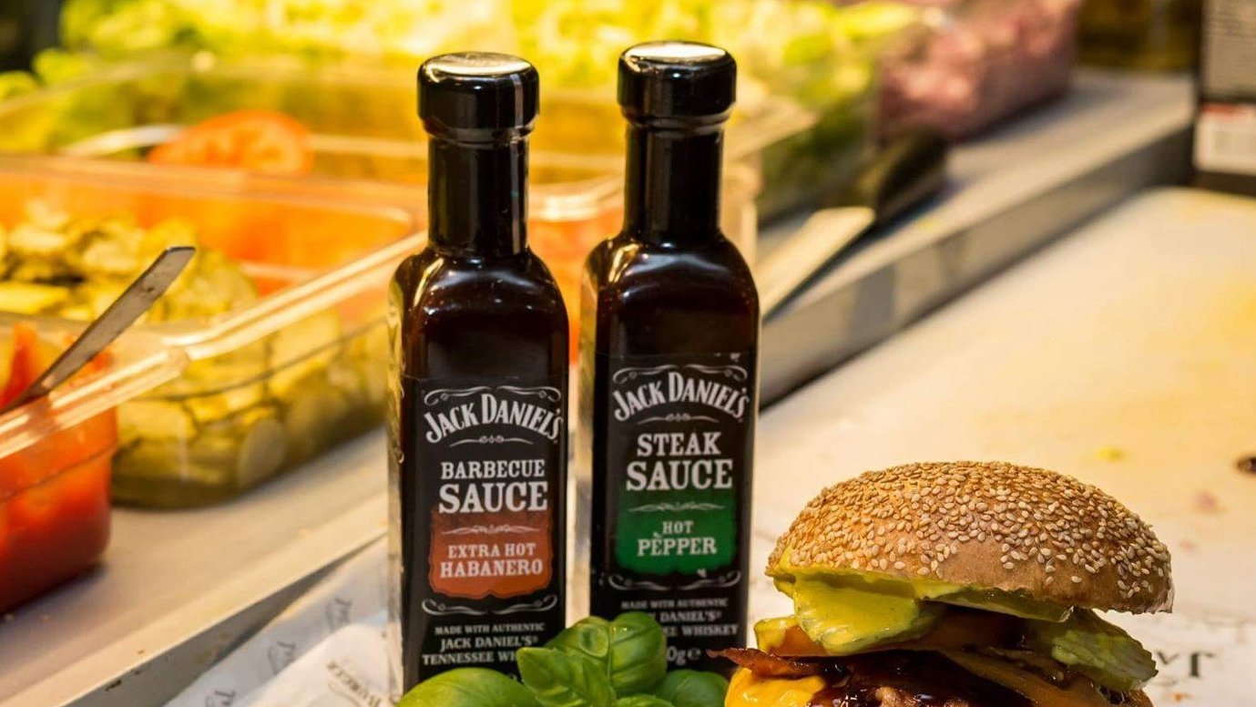 Duży burger z wieloma dodatkami leży na blacie. Obok burgera stoją dwie małe buteleczki z sosem "Jack Daniels". Z tyłu dodatki do burgera są umieszczone w pojemnikach.