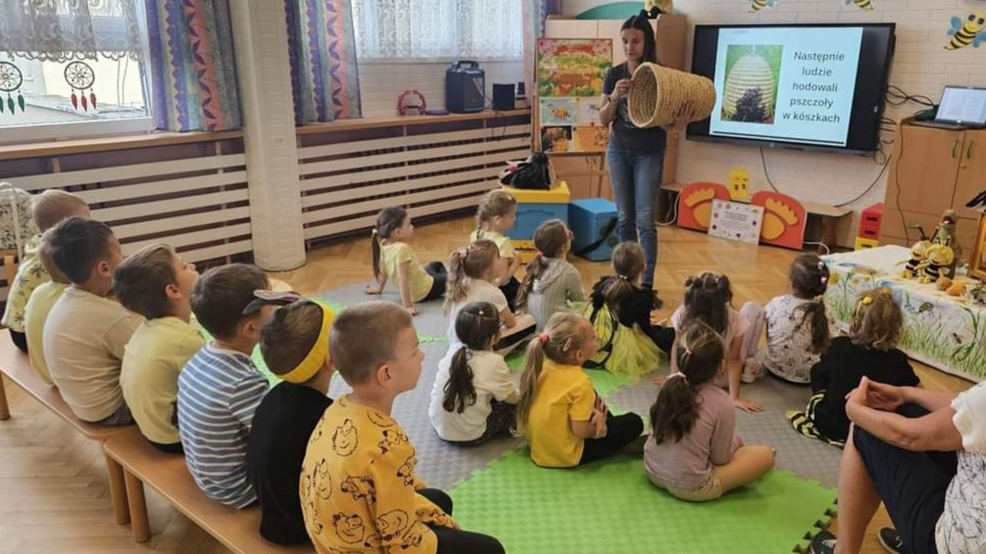 Zdjęcie przestawia siedzące na podłodze i ławeczkach dzieci słuchające wykładu o pszczołach.