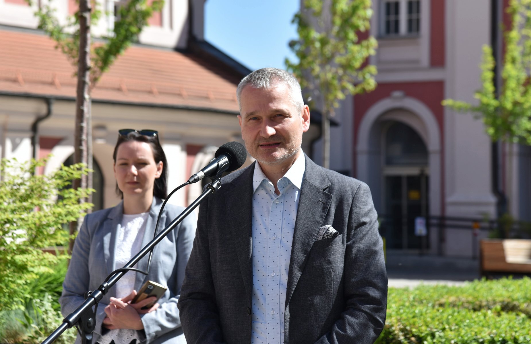 Na zdjęciu dwie osoby na dziedzińcu, mężczyzna przy mikrofonie, za nim kobieta - grafika artykułu