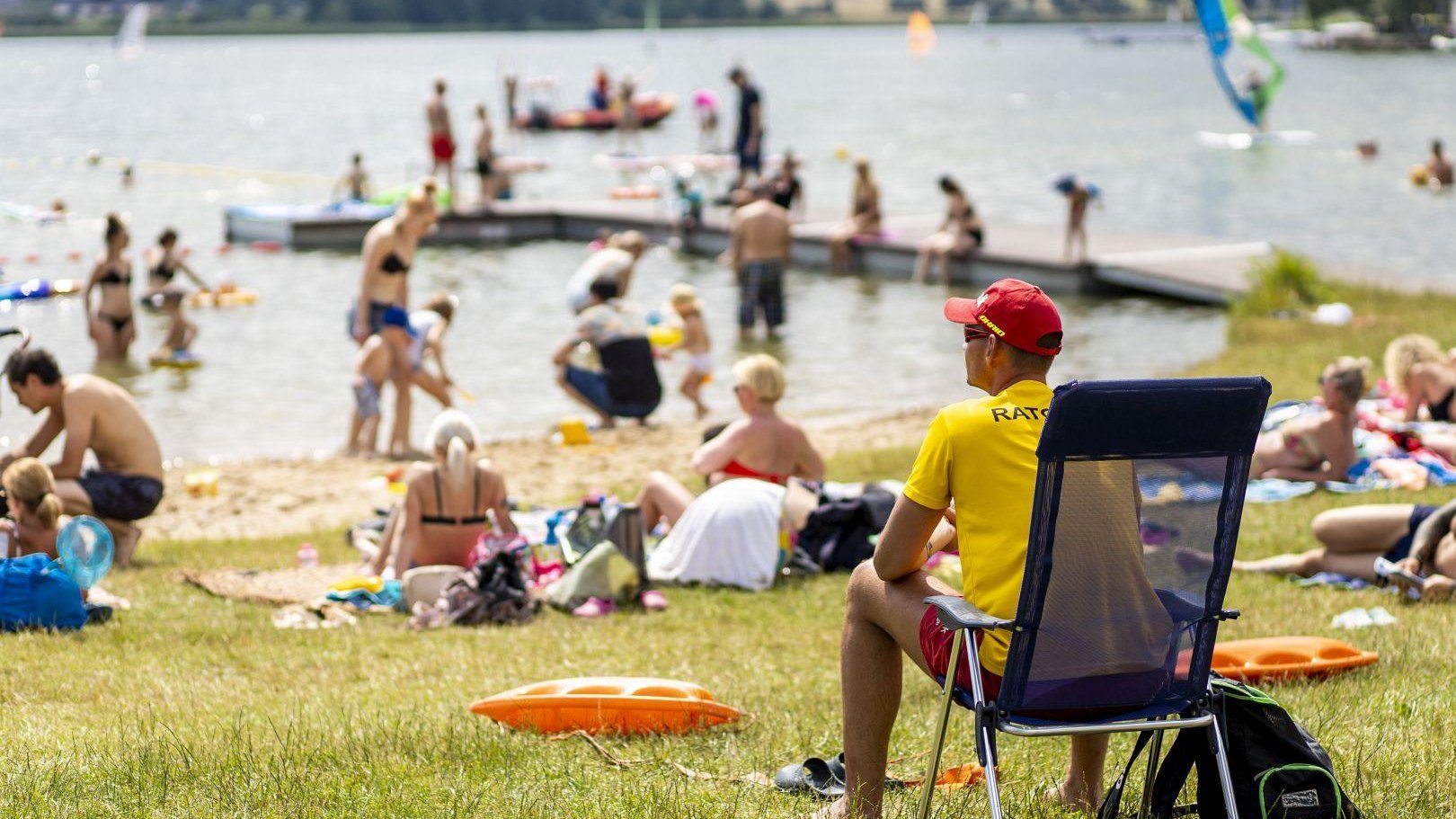 Zdjęcie przedstawia ratowanika siedzącego na plaży. W tle widać tłum ludzi na plaży i w wodzie.