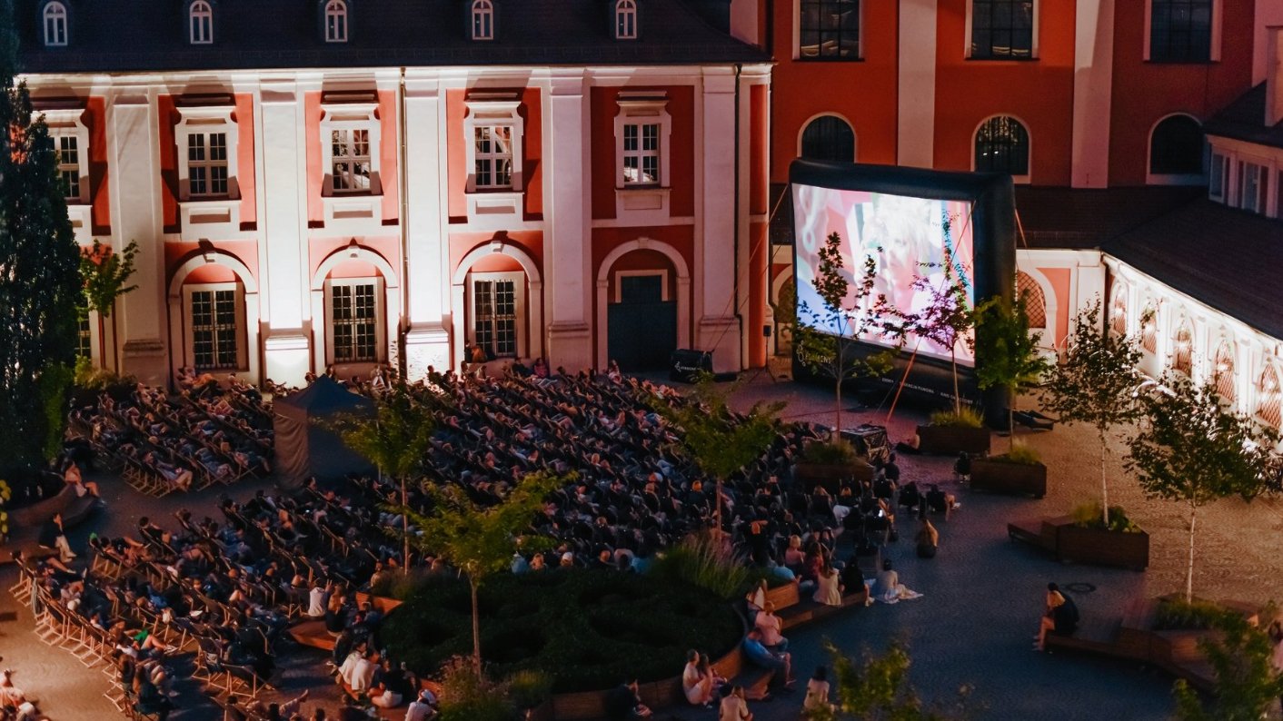 Zdjęcie przedstawia ludzi na leżakach podczas oglądania filmu na telebimie w plenerze.