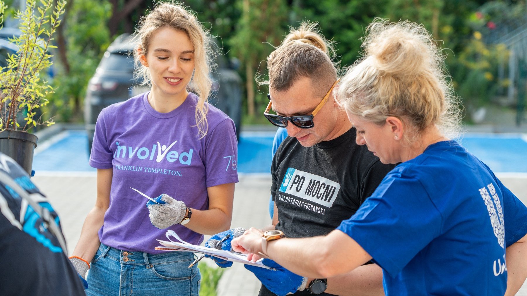 Na zdjęciu troje wolontariuszy pochylonych nad jakąś kartką, w otoczeniu zieleni