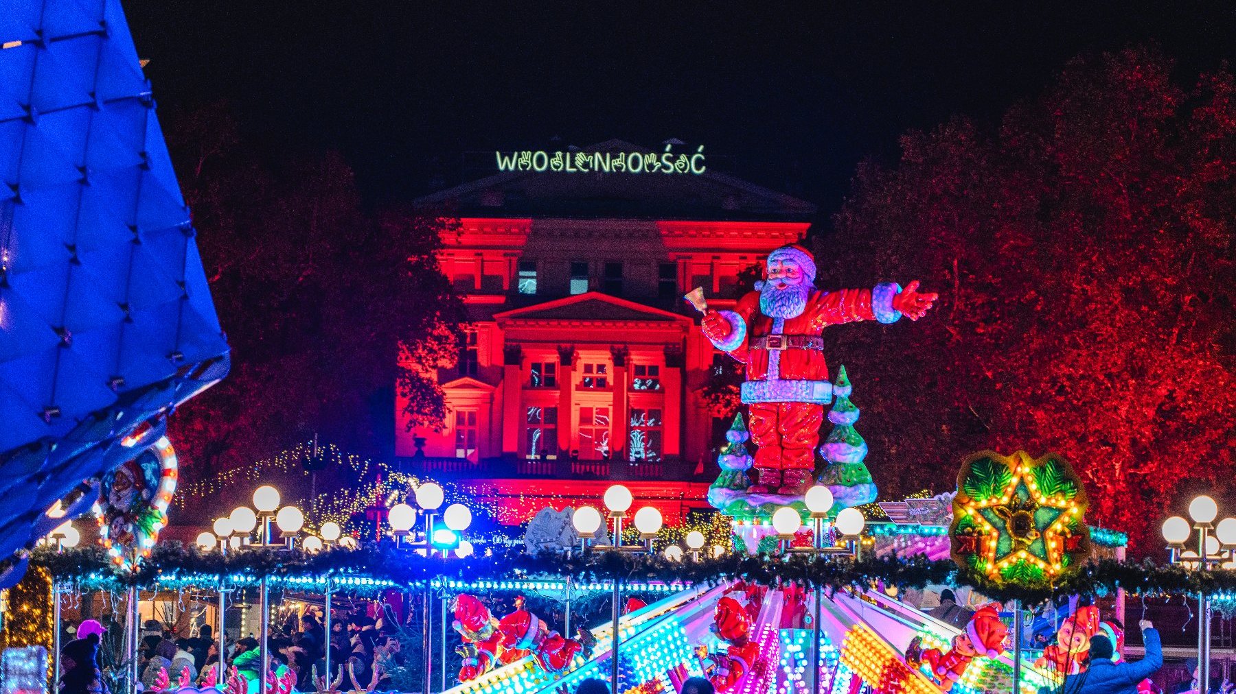 Zdjęcie przedstawia karuzelę ze Świętym Mikołajem, podświetlony budynek i tłum ludzi.