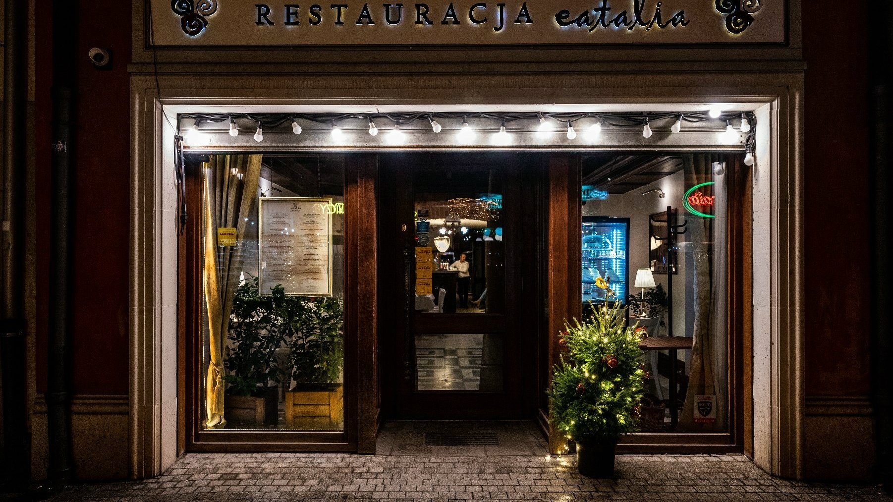 Zewnętrzna część restauracji o nazwie "Restauracja Eatalia". Wejście jest dobrze oświetlone, z zawieszonymi nad drzwiami lampkami. Po obu stronach drzwi znajdują się duże okna, przez które widać wnętrze restauracji. W lewym oknie znajduje się wywieszone menu, a przed oknami ustawione są rośliny. Po prawej stronie wejścia widoczna jest roślina w doniczce, ozdobiona lampkami. W środku restauracji widać stoły, krzesła, osobę oraz elementy dekoracyjne, takie jak lampa i neonowy znak. - grafika artykułu