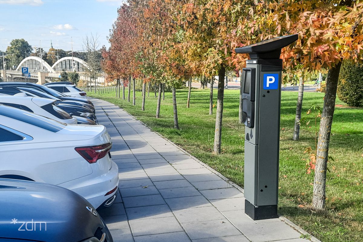 Zdjęcie parkomatu stojącego na chodniku pośród drzew z kolorowymi liśćmi. Po drugiej stronie chodnika rząd samochodów. W tle widac most św. Rocha - grafika artykułu