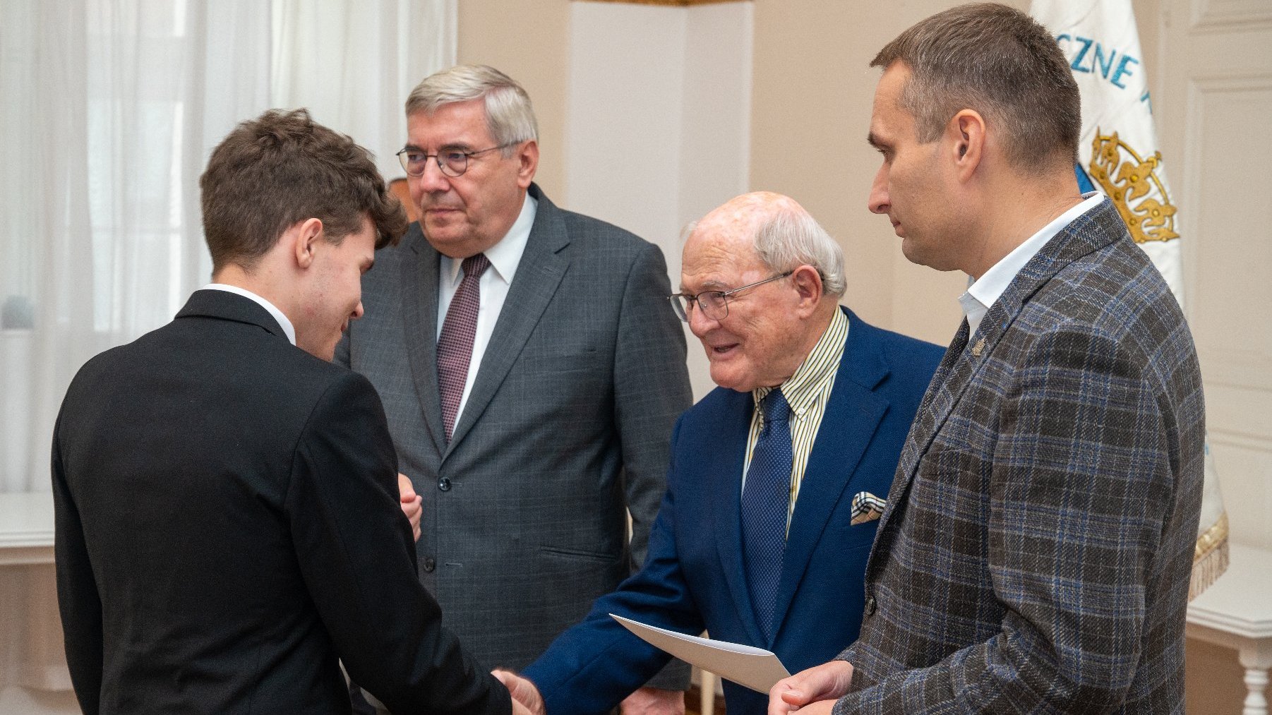 Zdjęcie przedstawia trójkę mężczyzn i młodego chłopaka podającego im rękę.