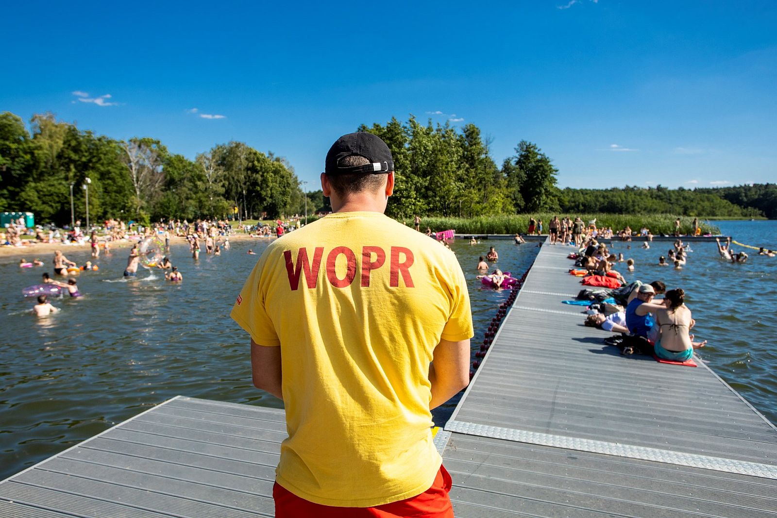 Ratownik WOPR w żółtej koszulce pilnuje bezpieczeństwa stojąc na drewnianym pomoście nad jeziorem, w wodzie kąpią się dzieci i młodzież - grafika artykułu
