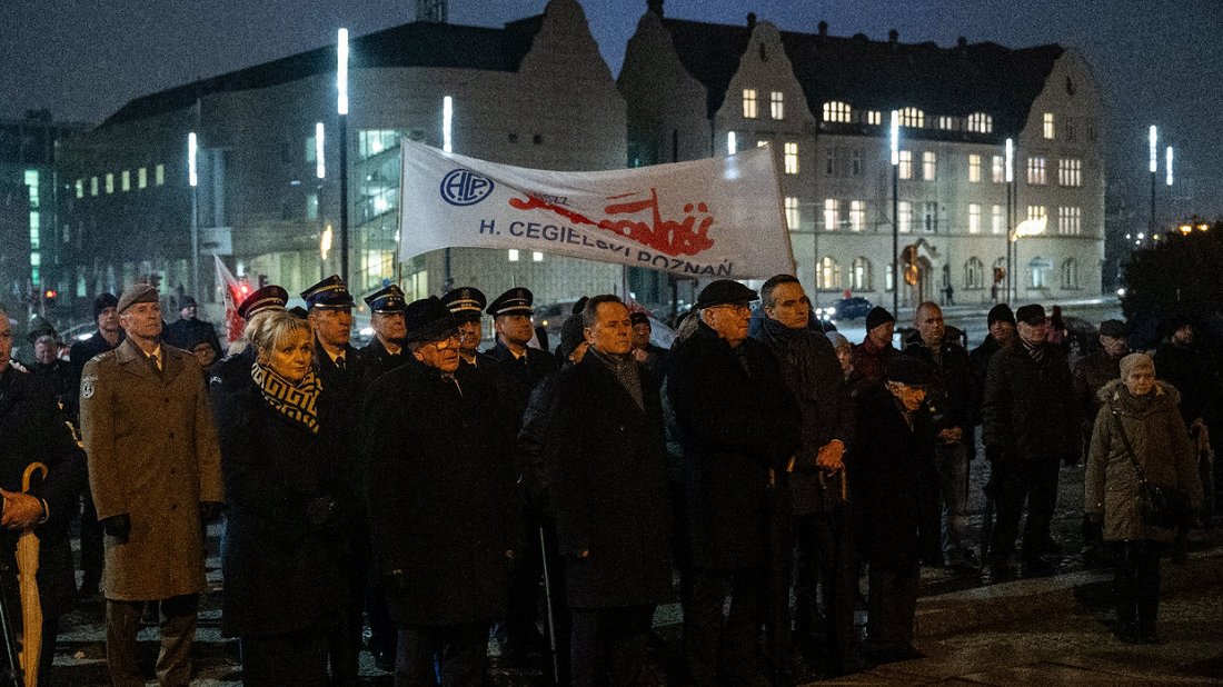 Grupa ludzi - kobiety i mężczyźni - w odzieniach zimowych stoi w szeregu są poażni, za nimi inna grupa ludzi trzyma transparent z napisem "Solidarność H. Cegielski Poznań", pora wieczorowa ciemni, w oddali światła budynków