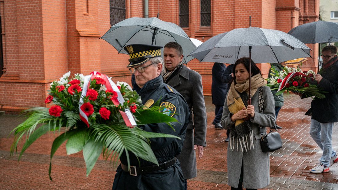 Na przedzie mężczyzna w mudurze Straży Miejskiej Poznania trzyma wiążankę kwiatów, za nim kobieta i mężczyzna trzymają parasole, w tle ceglany kościół