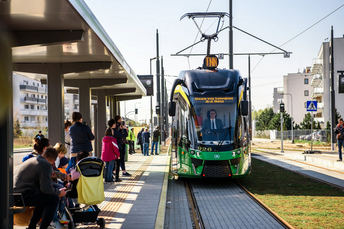 Tramwaj podjeżdża na przystanek, na nim wiaty. pod wiadami sporo osób w róznym wieku, za przystankiem i tramwajem widoczne bloki - grafika artykułu