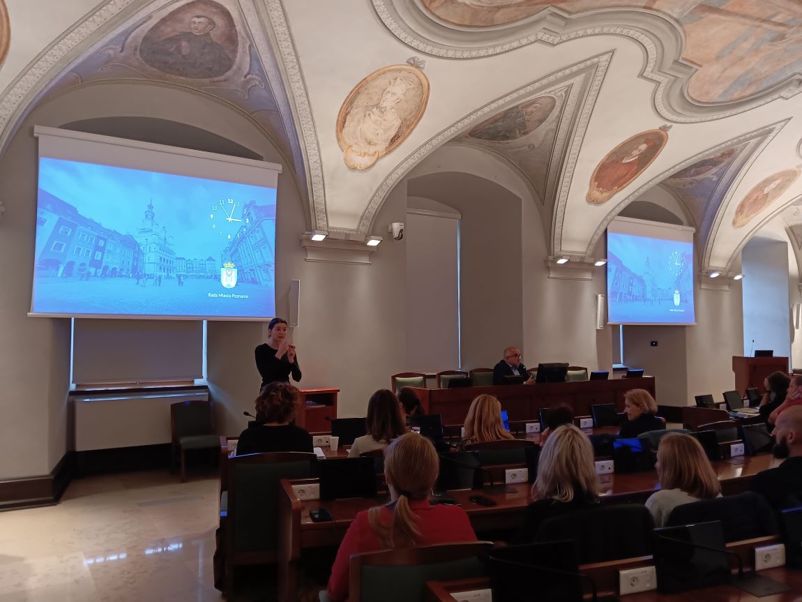 sala sesjna Rady Miasta Poznania, sklepienie zdobione freskami o tematyce sakralnej, za stanowiskami siedzą uczestnicy spotkania, przed nimi stoi tłumaczka języka migowego - grafika artykułu