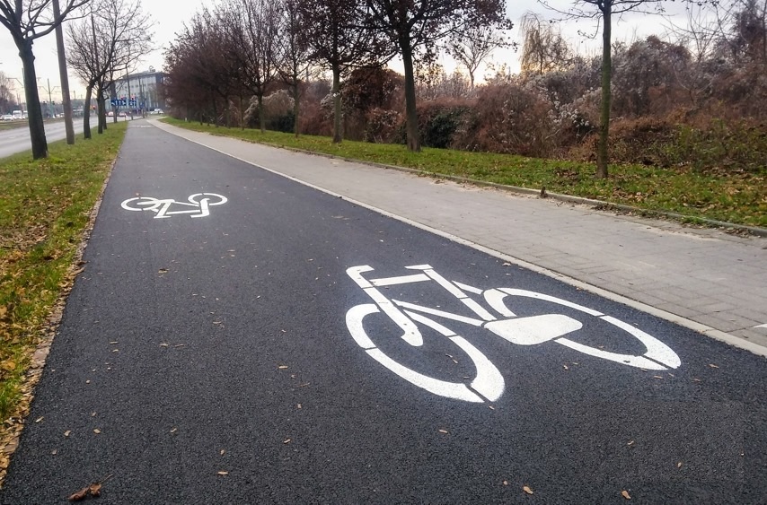 fgragmnet ścieżki rowerowej z wymalowanym symbolem roweru, obok chodnik, po obu stronach trawiaste pobocze i drzewa - grafika artykułu