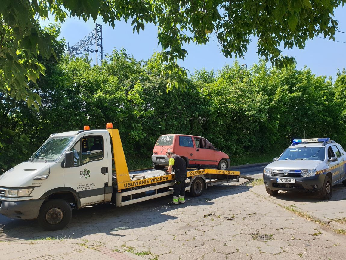 samochód laweta wciąga na lawetę samochód osobowy, człowiek przy lawecie obsługuje mechanizm, obok stoi samochód straży miejskiej, samochody stoją na asfalcie - grafika artykułu