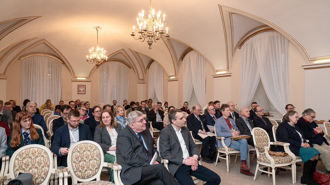 w sali ze zdobionymi filarami siedzą osoby nakrzesłach ustawionych w rzędy