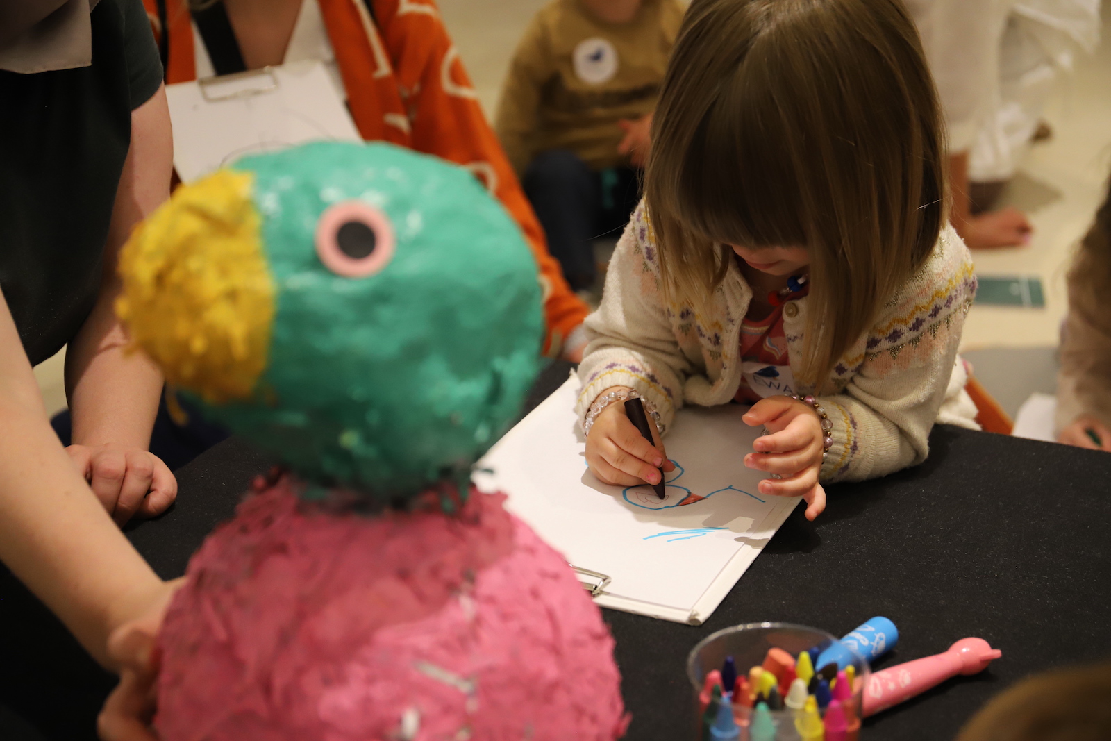 Zdjęcie, dziecko rysuje na kartce, przed dzieckiem stoi rzeźba kolorowego ptaka. - grafika artykułu