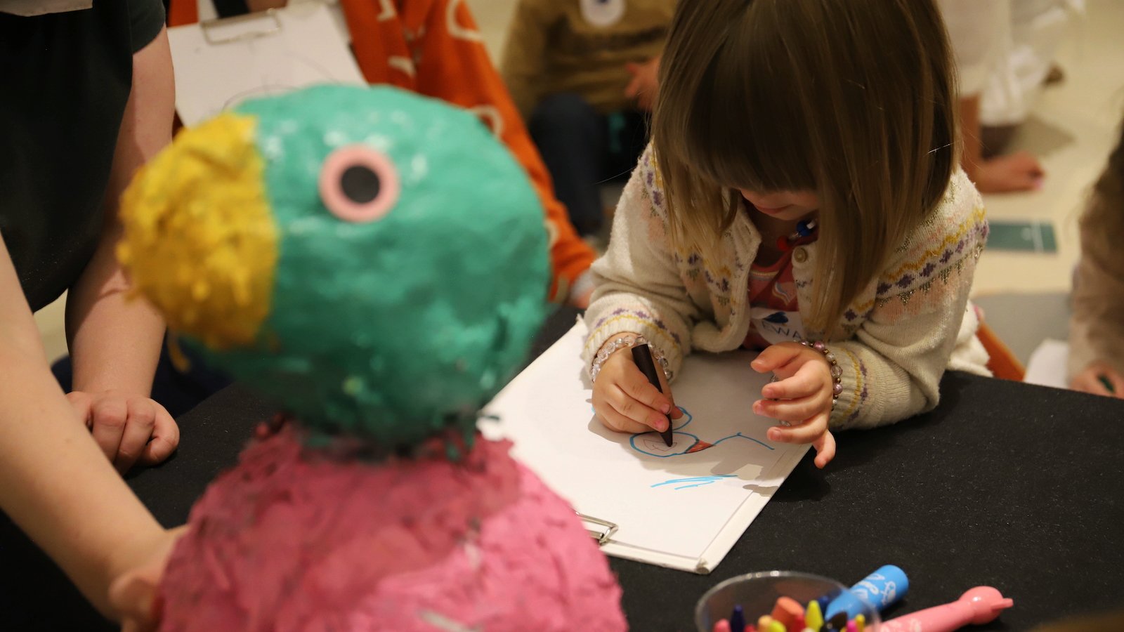 Zdjęcie, dziecko rysuje na kartce, przed dzieckiem stoi rzeźba kolorowego ptaka.