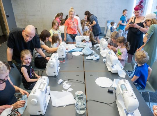 Zdjęcie, dzieci siedzą przy długim stole, na którym stoją maszyny do szycia, za dziećmi stoją dorości. - grafika artykułu