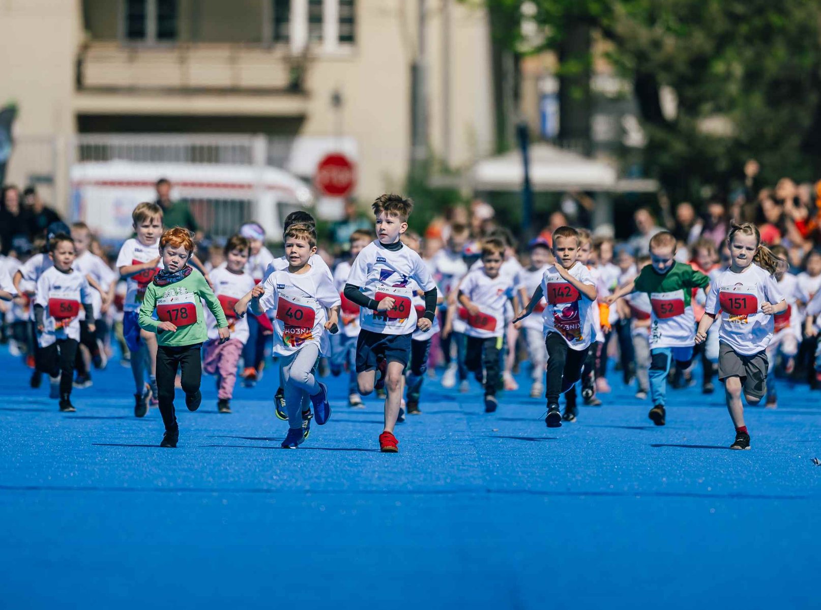 Grupa dzieci z numerami startowymi biegnie po niebieskiej nawierzchni. - grafika artykułu