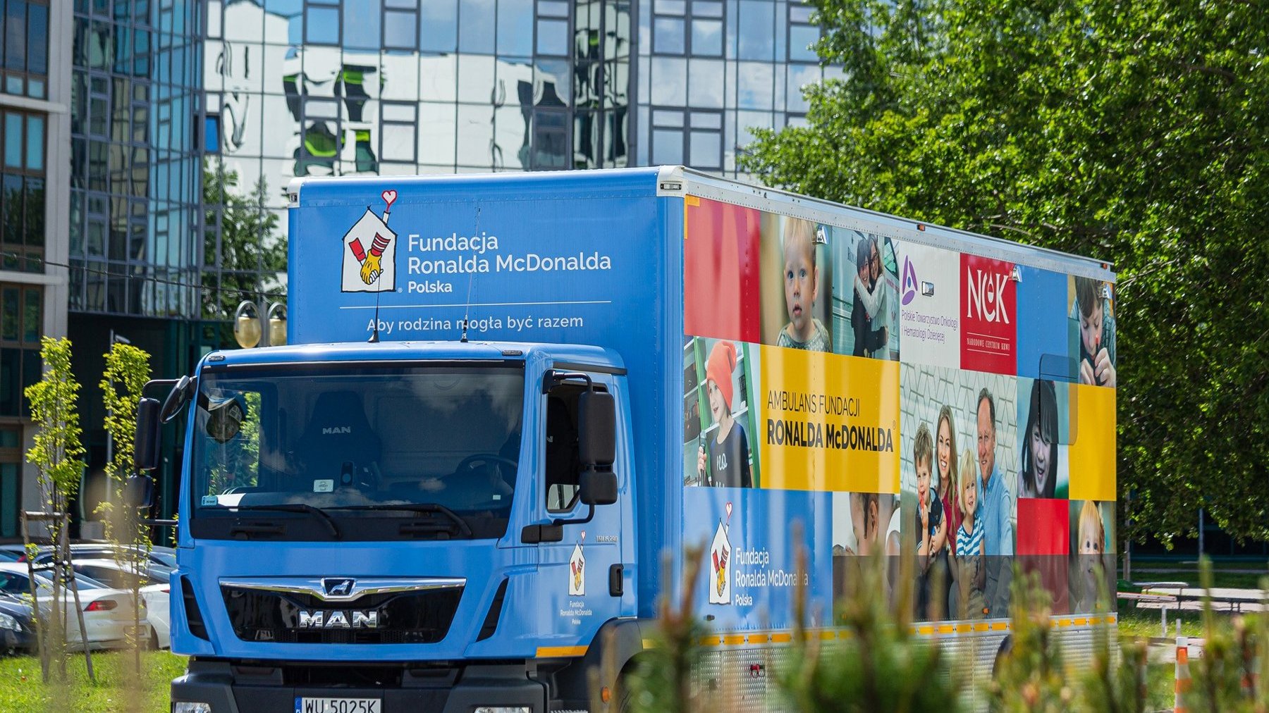 Zdjęcie przedstawia niebieską ciężarówkę Fundacji Ronalda McDonalda Polska, zaparkowaną w miejskim otoczeniu na tle nowoczesnych, przeszklonych budynków i zieleni. Na bocznej ścianie przyczepy widoczne są zdjęcia dzieci, rodzin oraz personelu medycznego, a także logo fundacji i innych partnerów. Na ciężarówce znajduje się napis: 'Aby rodzina mogła być razem' oraz oznaczenia sugerujące, że pojazd pełni funkcję ambulansu fundacyjnego.