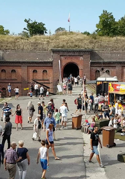 Tłumy ludzi w letnich strojach chodzących na dziedzincu Fortu VI. Na nim znajduja się zstragany z jedzeniem, pamiątkami i zabawkami.