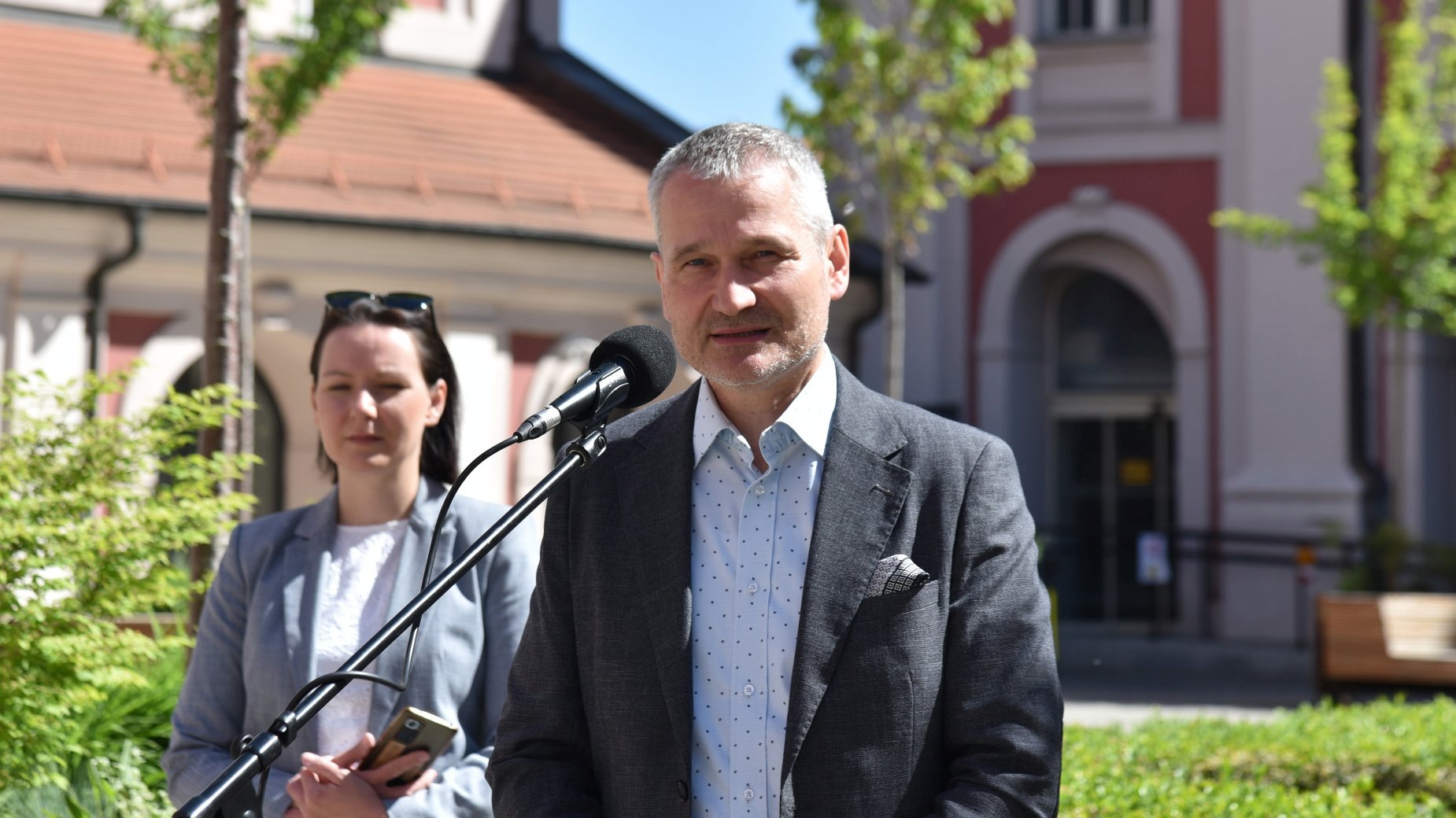 Na zdjęciu dwie osoby na dziedzińcu, mężczyzna przy mikrofonie, za nim kobieta