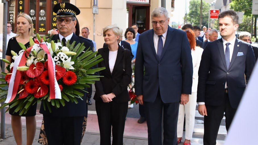 Na zdjęciu kilka osób składających wiązankę kwiatów, na pierwszym planie strażnik miejski z kwiatami