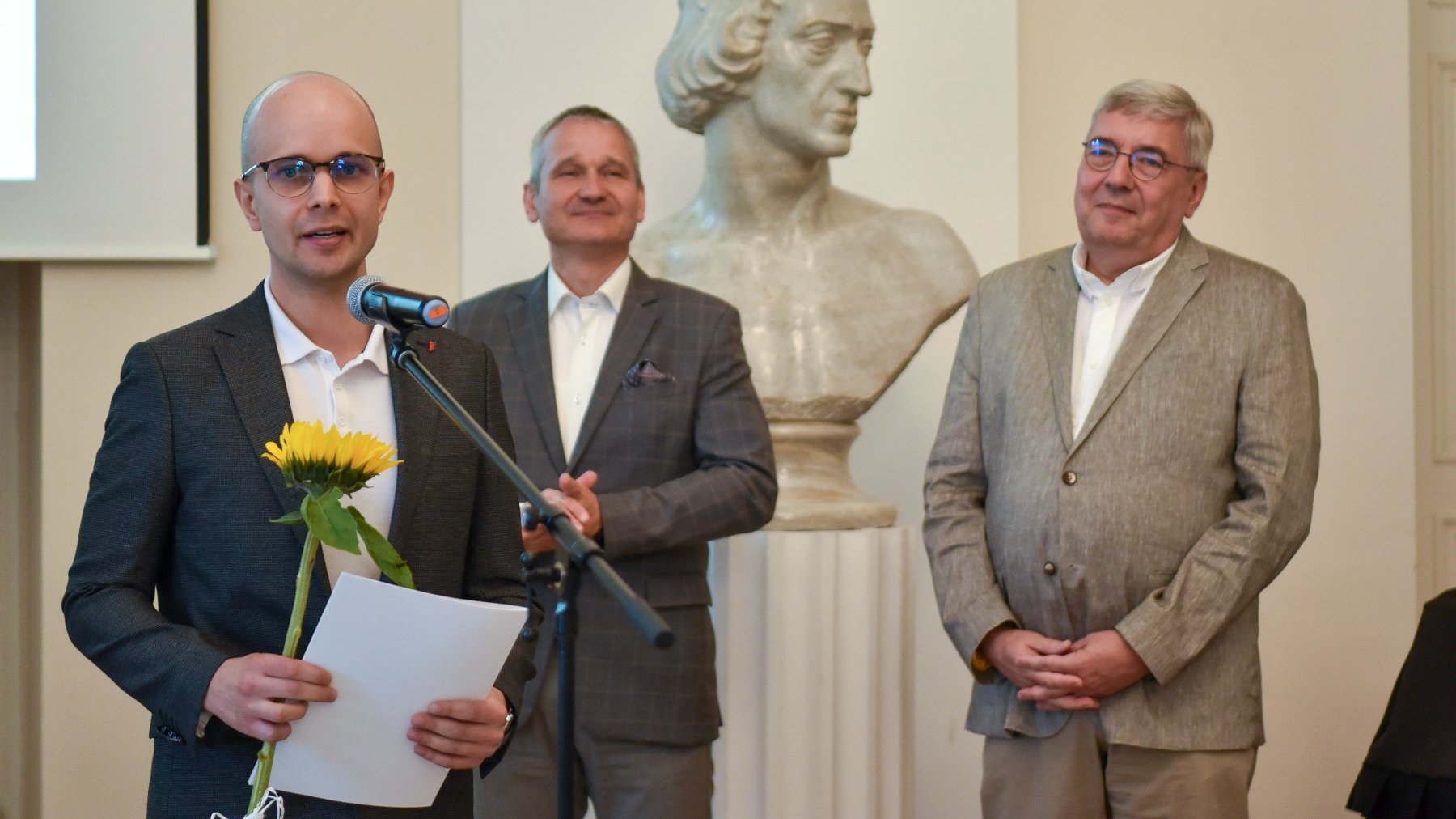 Na zdjęciu młody mężczyzna w okularach mówi coś do mikrofonu, za nim dwóch starszych mężczyzn