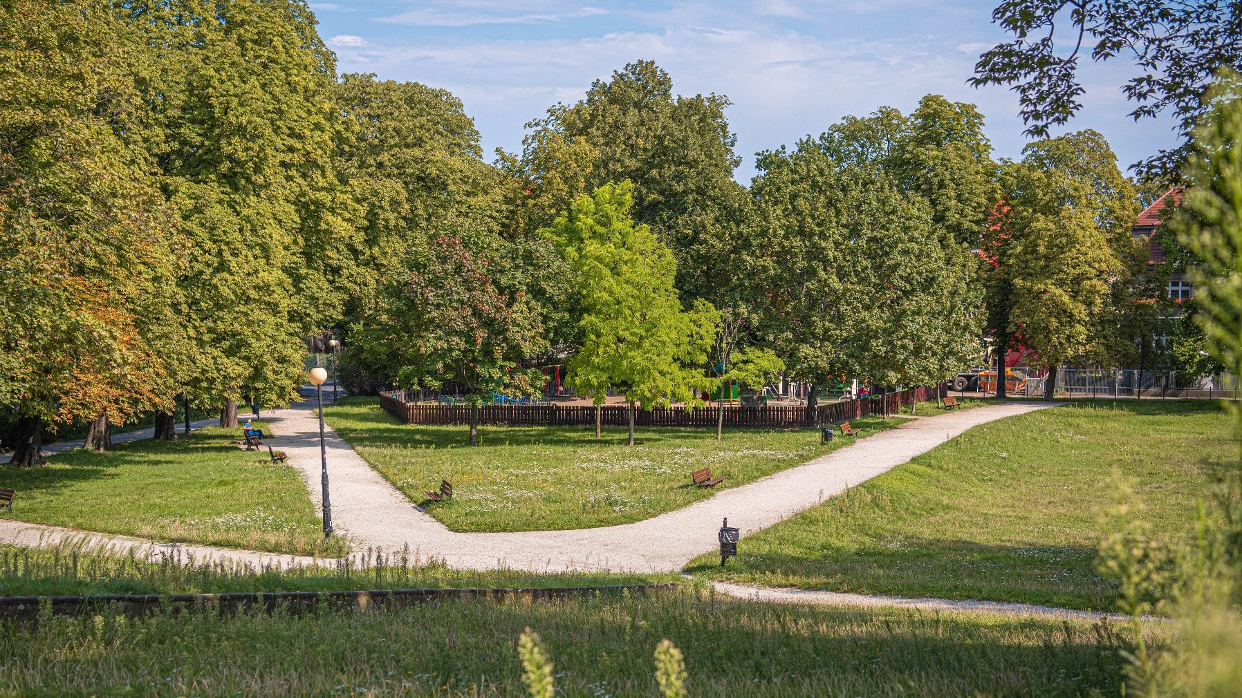 Zdjęcie przedstawia park. Widać na nim zieleń - trawę i drzewa oraz alejki z ławkami. W tle znajduje się ogrodzenie, za którym jest plac zabaw.