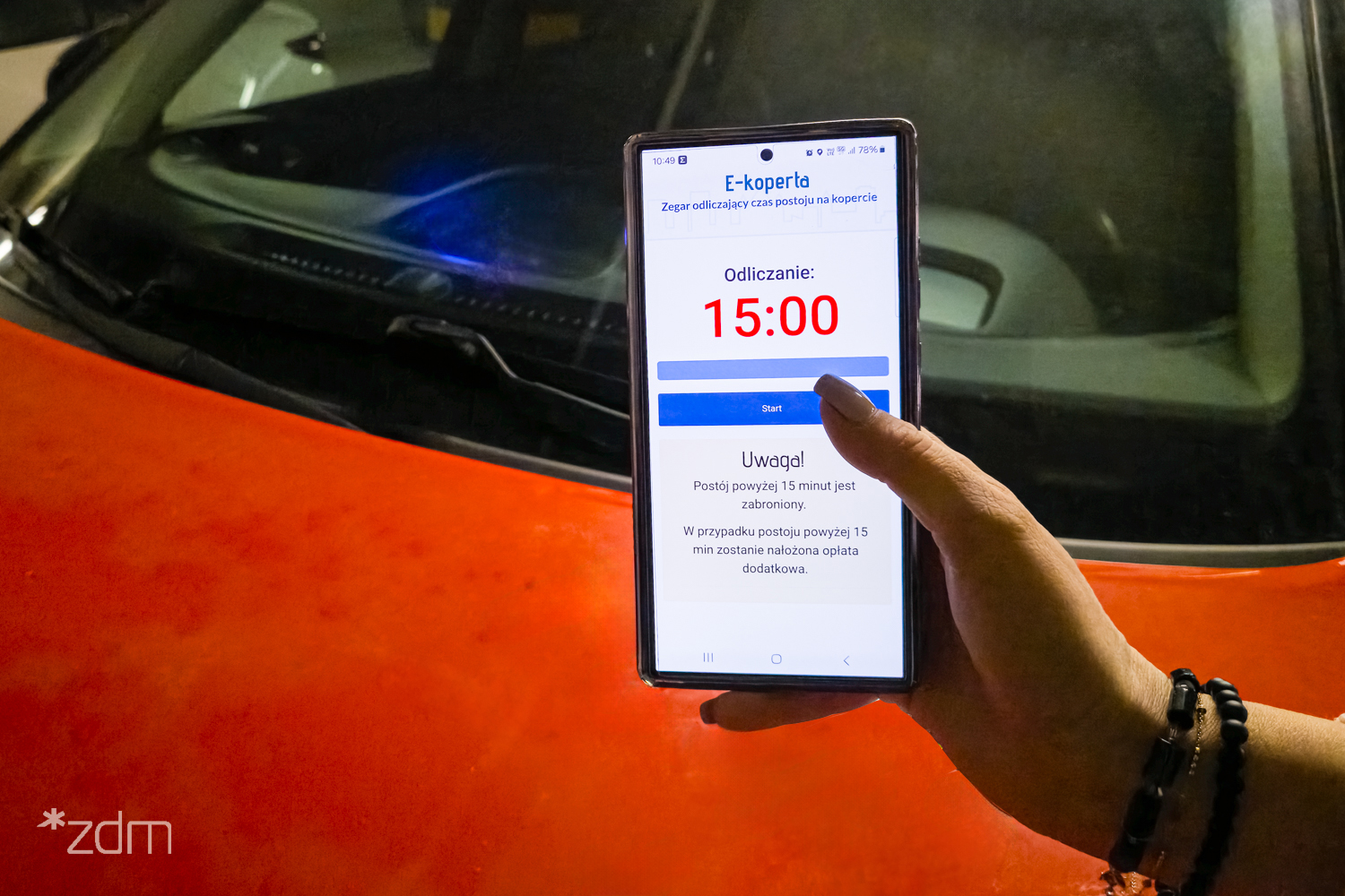 Kobieca ręka trzyma telefon z elektronicznym zegarem odmierzającym czas parkowania. W tle pomarańczowy samochód - grafika artykułu