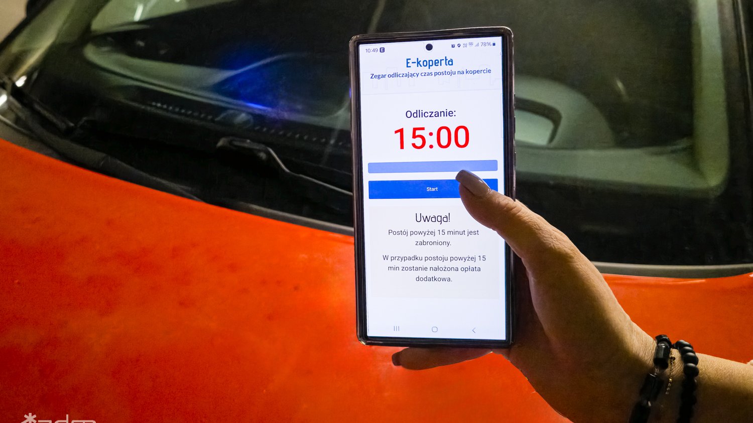 Kobieca ręka trzyma telefon z elektronicznym zegarem odmierzającym czas parkowania. W tle pomarańczowy samochód