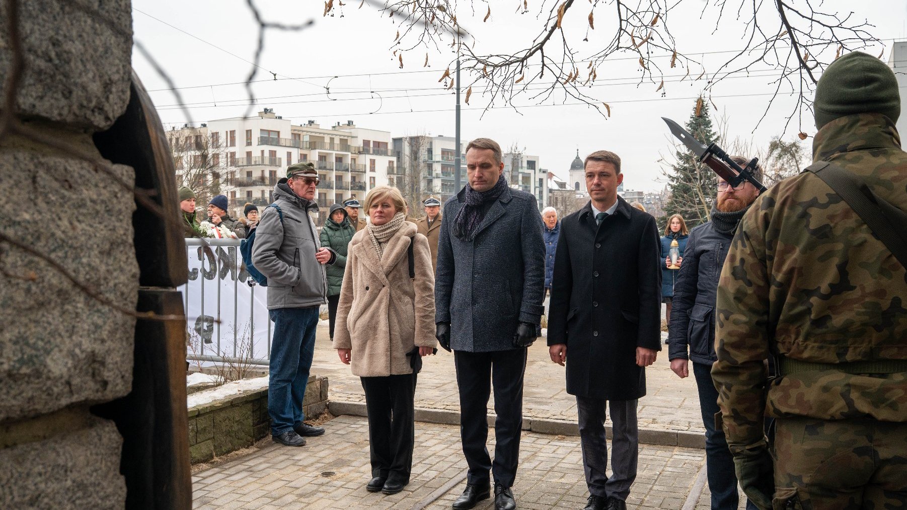 Grupa osób stoi na zewnątrz na utwardzonym terenie. Na pierwszym planie widzimy czterech ludzi ustawionych w szeregu, zwróconych w stronę osoby w mundurze kamuflażowym, która trzyma duży nóż. W tle znajdują się inne osoby, z których niektóre trzymają bannery, oraz rząd nowoczesnych bloków mieszkalnych.