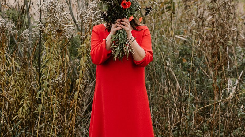 Kobieta w czerwonej sukience stoi boso w wysokiej trawie. W rękach trzyma bukiet z czerwonymi kwiatami, którymi zasłania swoją twarz. Za nią wysokie trawy.