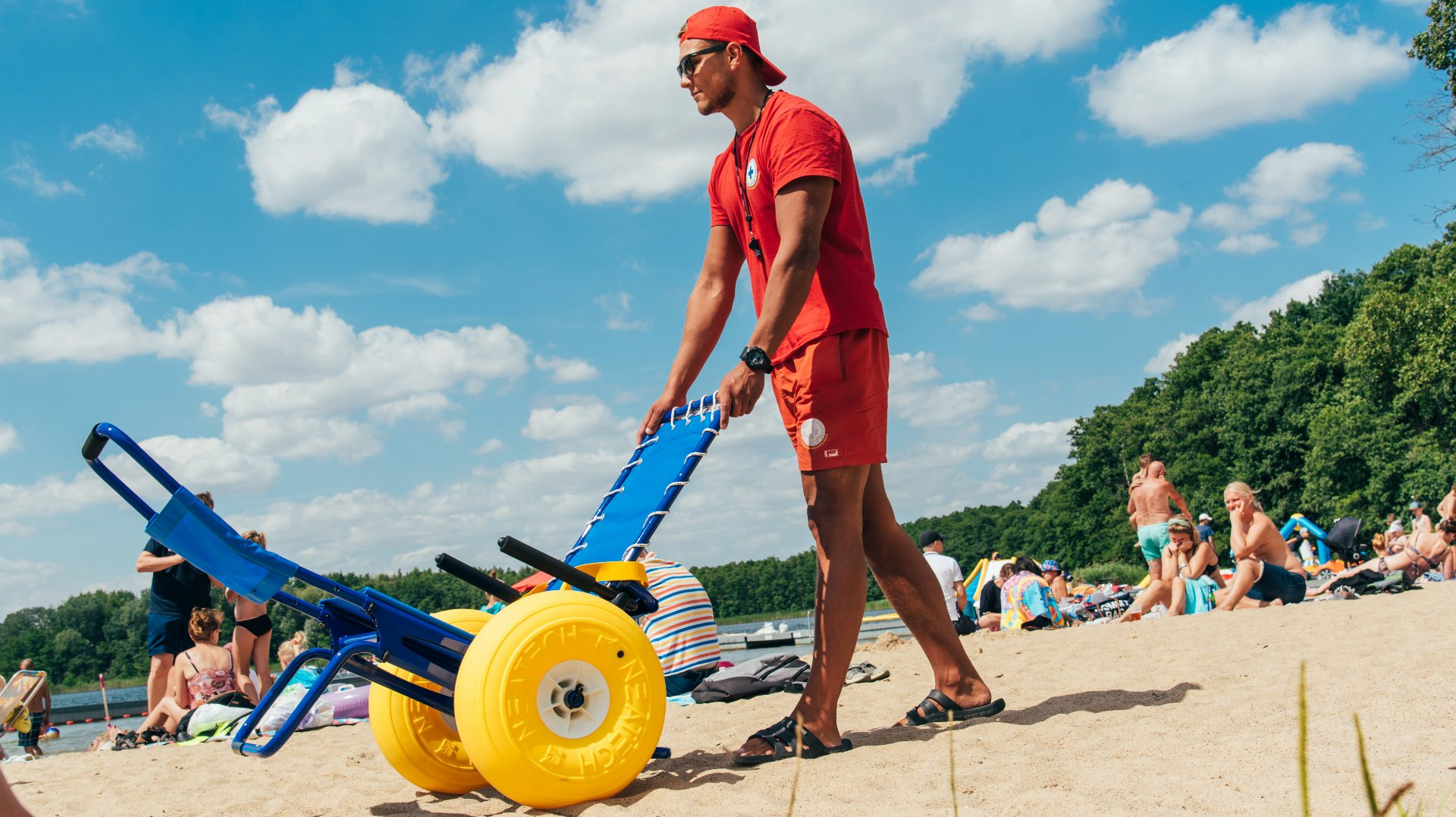 Mężczyzna, który jest ratownikiem wodnym trzyma wózek-amfibię, prowadzi go na plaży - grafika artykułu