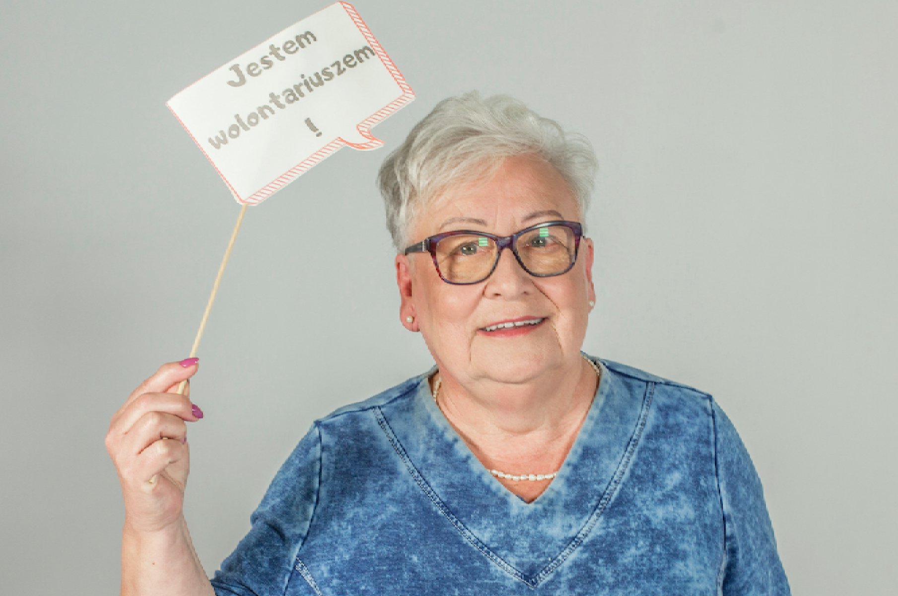 starsza pani trzymająca tabliczkę nad głową z napisem - jestem wolontariuszem - grafika artykułu