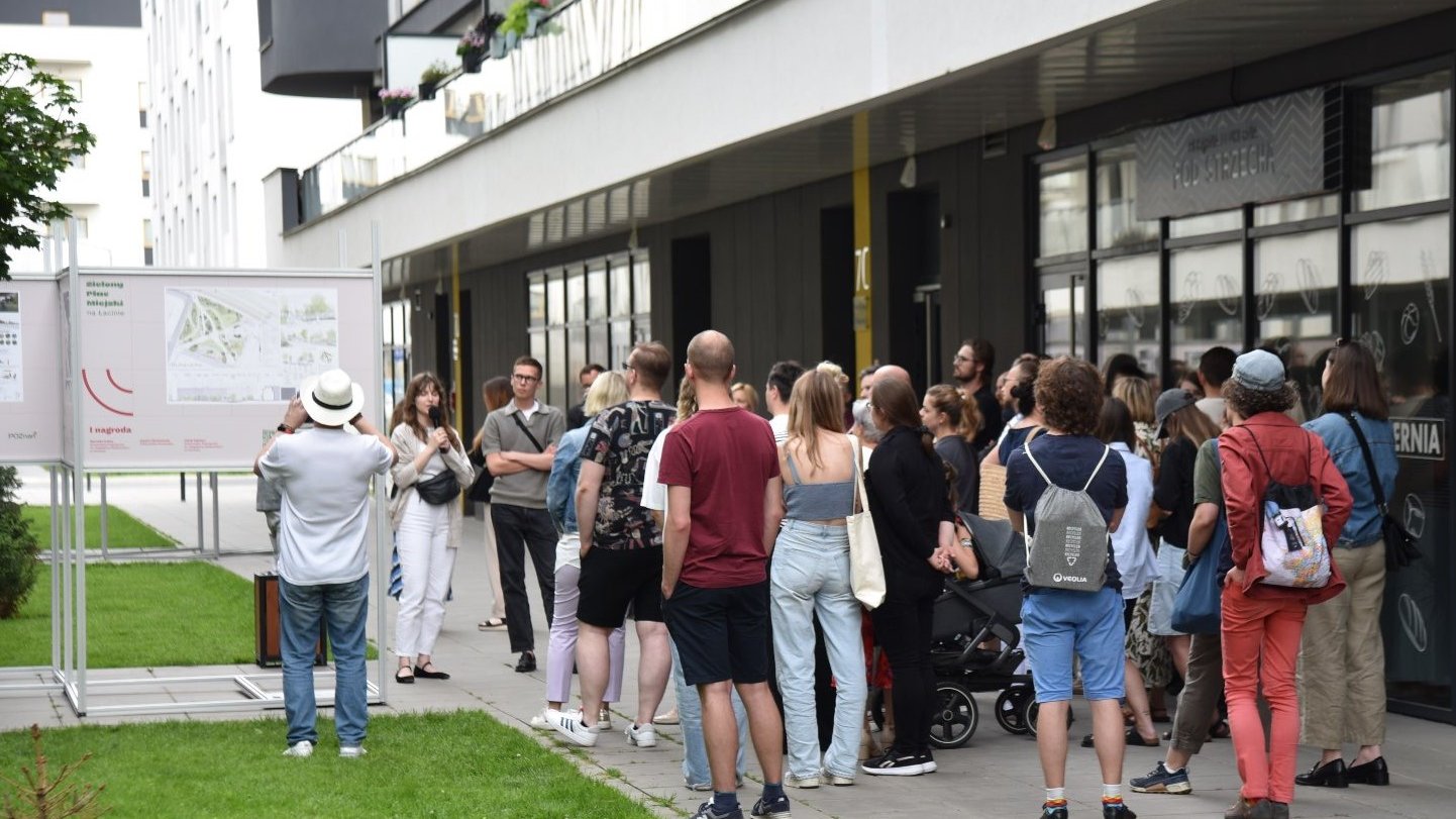 Grupa osób stoi przy nowoczesnym budynku, na chodniku obok trawnika.