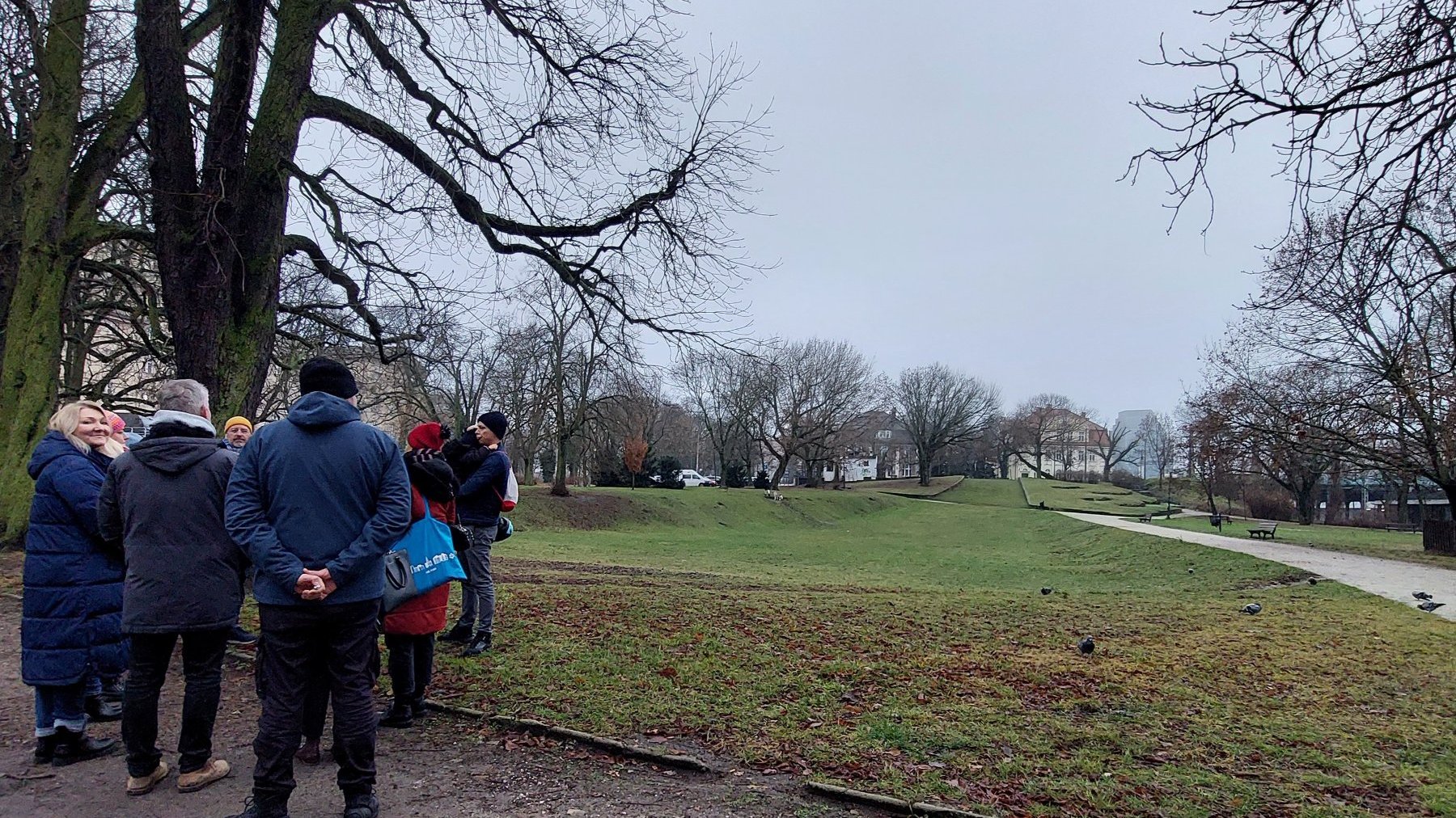 Kilka osób stoi przy parkowej drodze, w tle park i oddalone budynki.