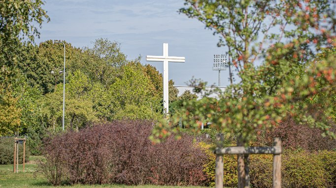 Biały krzyż na lekkim wzniesieniu w parku, otoczony zielelenią.