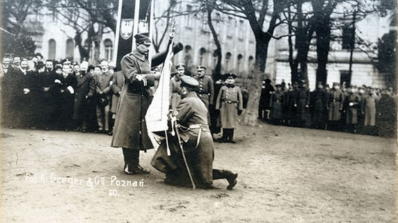 Zołnierz klęczący przed innym żołnierzem, ze sztandarem w ręku. W tle żołnierze i cywile.