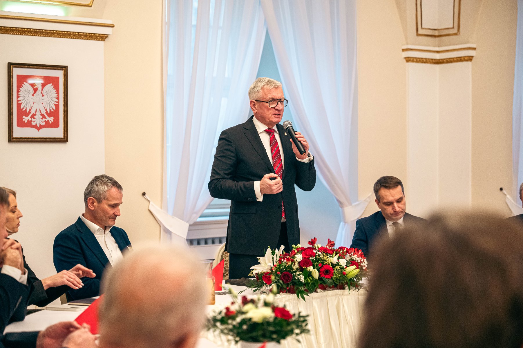 Zdjęcie przedstawia formalne spotkanie lub konferencję, na której siedzą kilka osób wokół stołu. Centralna postać stoi i mówi przez mikrofon, ubrana w garnitur i krawat w czerwone i białe paski. Stołek ozdobiony jest kwiatami, a na ścianie znajduje się wizerunek białego orła na czerwonym tle, który jest często kojarzony z Polską. - grafika artykułu