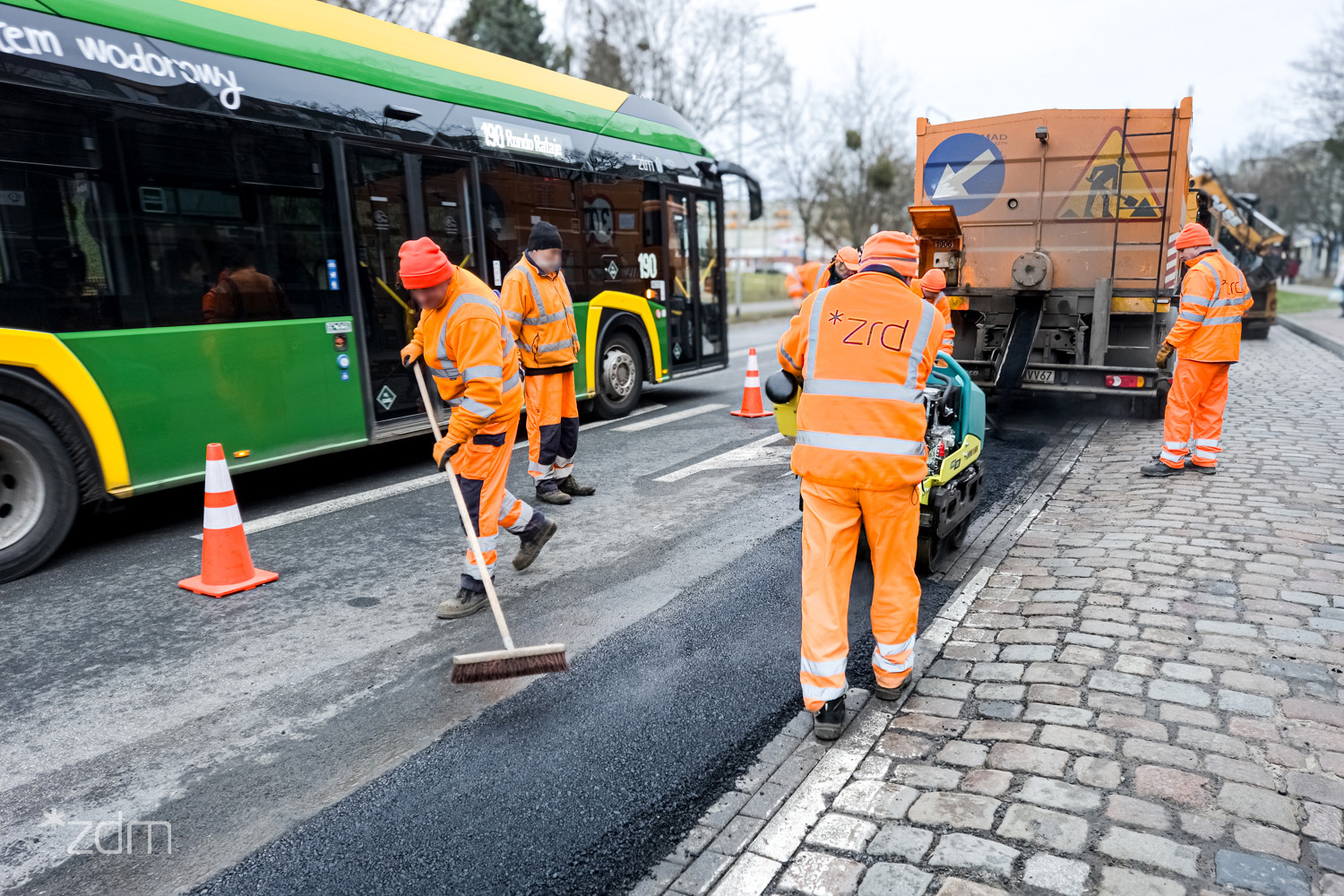 Ekipa pracowników Zakładu Robót Drogowych naprawia jezdnię. Obok widać przejeżdżający autobus - grafika artykułu