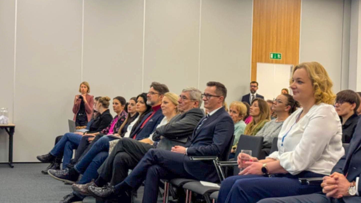 Grupa ludzi siedzących w sali konferencyjnej. Osoby siedzą w rzędach krzeseł, zwrócone w lewo, co sugeruje, że słuchają prelekcji lub prezentacji. Sala ma nowoczesny design, białe ściany, drewniany panel oraz sufit z wbudowanym oświetleniem. W tle stoi osoba, prawdopodobnie ochroniarz lub członek obsługi wydarzenia.