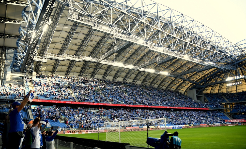 Na zdjęciu jedna z trybun Stadionu Miejskiego w Poznaniu podczas meczu Lecha Poznań. Na trybunie widać wielu wiwatujących kibiców, ubranych w niebiesko-białe barwy Lecha. Na pierwszym planie zielona murwa stadionu i jedna z bramek piłkarskich - grafika artykułu